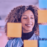 A imagem mostra uma mulher de frente para um quadro com post-its nas cores amarelo e azul. A foto sugere um processo de planejamento de go-to-market.