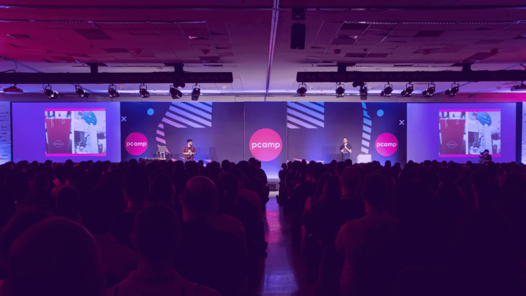 A imagem mostra o palco e a plateia do Product Camp em 2019, reforçando que o Product Camp 2022 vai retomar os encontros presenciais.