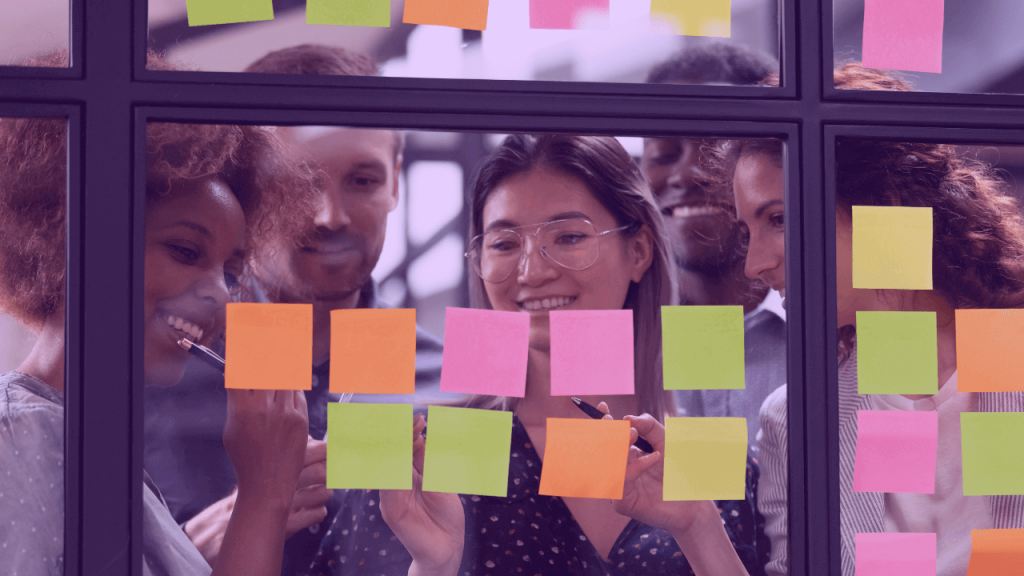 A imagem mostra um grupo de pessoas sorrindo atrás de um vidro com vários posti-its coloridos. A foto sugere que o grupo está aplicando a dinâmica 10 Star Experience and Beyound para descobrir a melhor experiência para os usuários de seu produto.