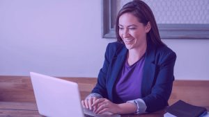 A imagem mostra uma mulher sorrindo para o computador. A foto sugere que ela está se preparando para uma migração de carreira para Produto.